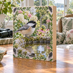 Black-capped chickadee wooden block perched on a mossy birdbath with reflected leaves in a botanical sunroom.
